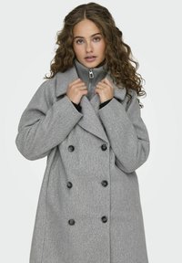 Cappotto doppio petto in misto lana grigia con grandi bottoni neri, dotato di colletto a rever e un maglione grigio con zip e colletto alto sotto.