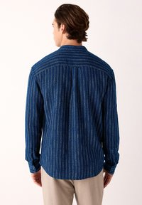 Man met bruin haar, gekleed in een blauw, lang mouwen, gebloemd overhemd en beige broek, staand met de rug naar de kijker tegen een effen achtergrond.