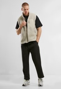 Beige fleece vest with a high collar, front zip, and one chest pocket; paired with a black shirt and black pants, standing pose.
