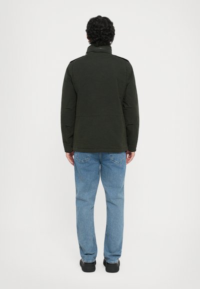 Dunkelgrüne Jacke mit hohem Kragen, aus leichtem Stoff gefertigt. Kombiniert mit hellblauen Jeans und schwarzen Schuhen, von hinten betrachtet.