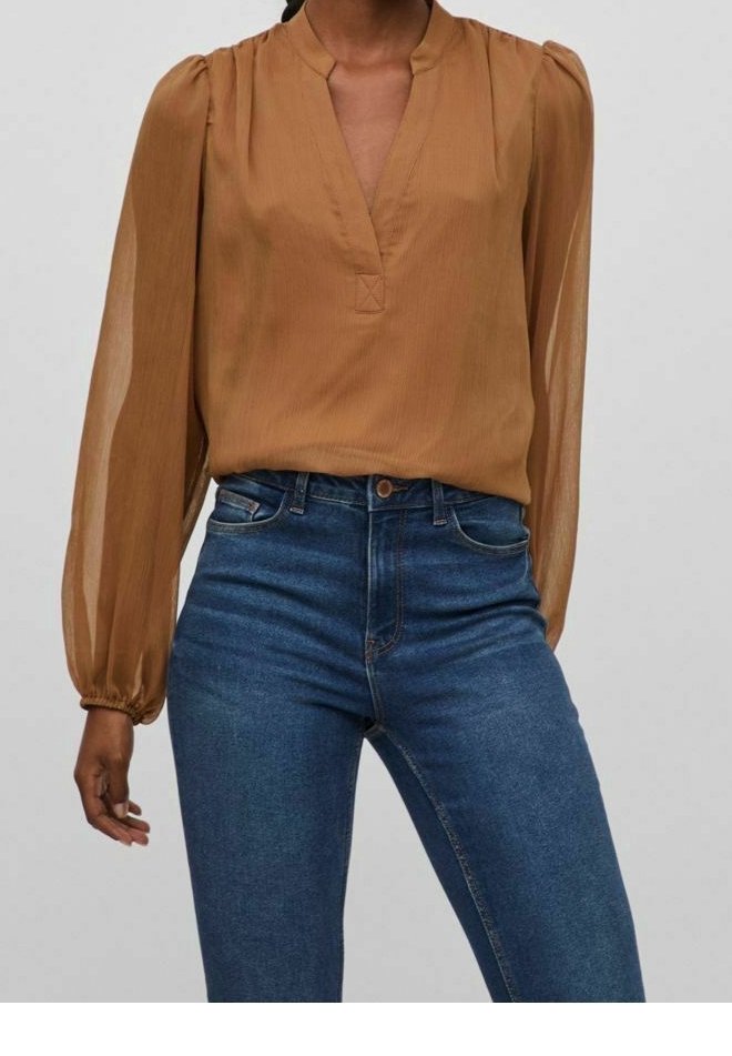 Femme portant un chemisier translucide manches longues couleur rouille avec un col en V et un jean bleu taille haute, sur un fond uni.