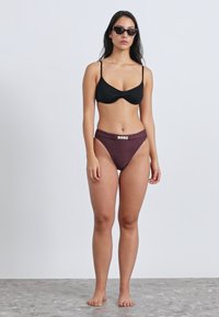 Zwarte bikinibovenkant met verstelbare bandjes, gecombineerd met bordeauxrode high-waisted broekjes met een decoratief gouden accent. Soepele stoftextuur.