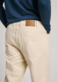 Pantalones beige con una textura suave, que presentan dos bolsillos traseros y un parche de cuero marrón claro en la cinturilla. Combinados con una camisa de manga larga azul marino.