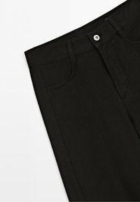 Close-up af sorte denimjeans med synligt linning, knaplukning, bæltestropper og frontlomme mod en hvid baggrund.