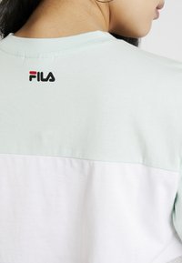 T-shirt en coton vert menthe et blanc à manches courtes, avec un logo FILA en noir et rouge au dos. Texture lisse, style décontracté.