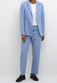 Completo blu chiaro con blazer a un bottone e pantaloni su misura, caratterizzato da una texture morbida e un design minimalista. Abbinato a una t-shirt bianca.