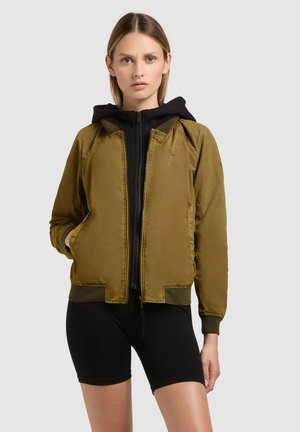 Ung kvinde iført en olivengrøn bomberjakke over en sort lynlåshoodie og sorte cykelshorts, stående foran en ensartet baggrund.