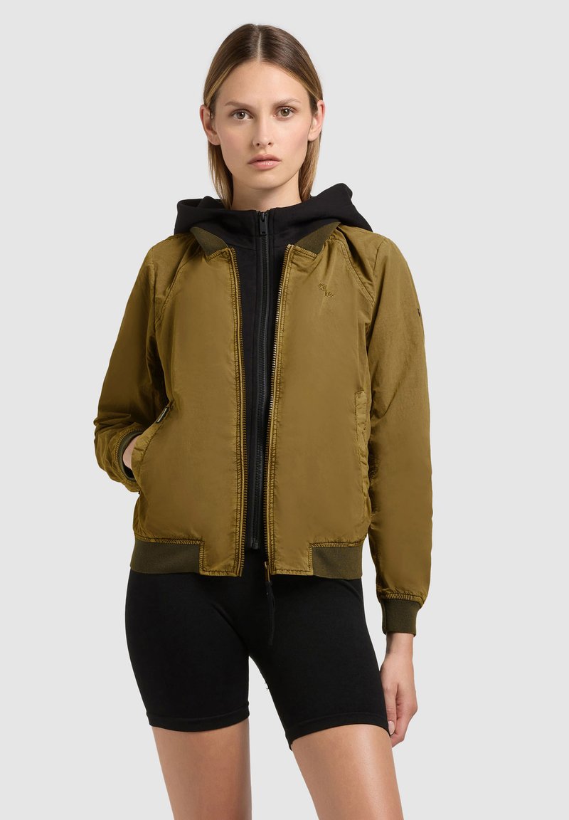 Junge Frau trägt eine olivgrüne Bomberjacke über einem schwarzen Kapuzenpullover mit Reißverschluss und schwarzen Radsport-Shorts, sie steht vor einem einfarbigen Hintergrund.