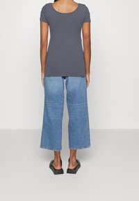 Mujer de pie de espaldas, vestida con una camiseta gris de manga corta, jeans azules de pierna ancha y tobillo, y sandalias de plataforma negra con talones abiertos.