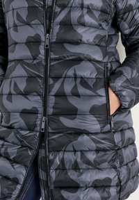 Zwarte en grijze gewatteerde jas met een camouflagepatroon, voorzien van horizontale naden, ritsen en zijzakken. Zachte, doorgestikte textuur.