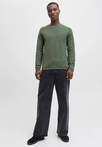 Pull vert à manches longues avec un motif texturé, associé à un jean noir ample et des chaussures foncées, sur fond clair.