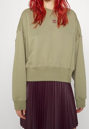 Persona con pelo rojo lleva una sudadera Adidas verde oliva y una falda plisada de color púrpura oscuro, mostrada desde los hombros hasta la mitad del muslo.