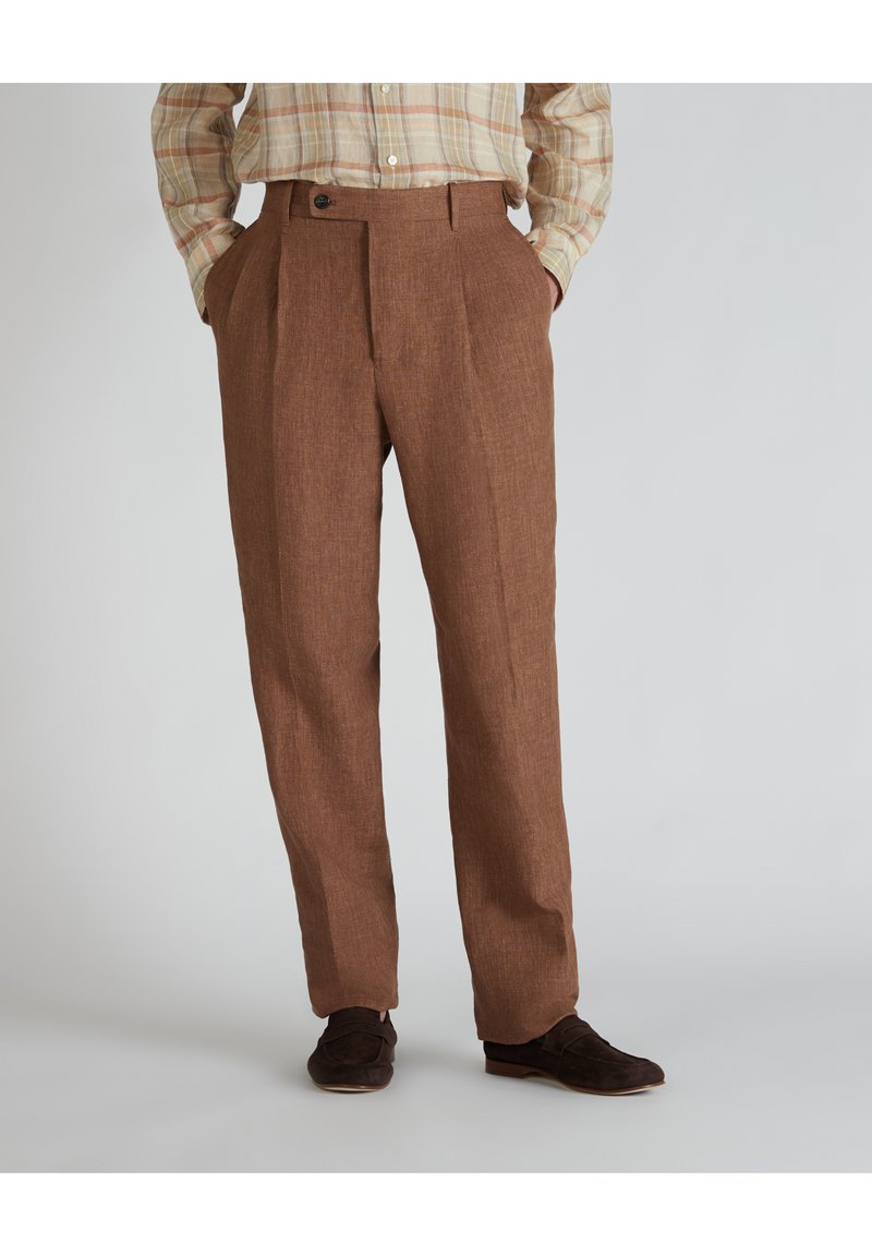Pantaloni in lino marrone con una finitura testurizzata, dotati di pieghe frontali e chiusura con un solo bottone. Abbinati a scarpe scure.