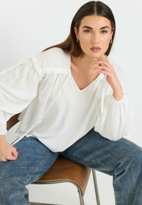 Hvid bluse med voluminøse ærmer og sammenkrøllede detaljer, parret med blå denimjeans. Stoffet ser letvægts ud med en glat tekstur.