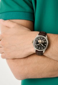 Reloj con correa de cuero marrón, caja redonda de plata, esfera negra, marcadores blancos y dos subesferas. La correa tiene perforaciones para un aspecto texturizado.