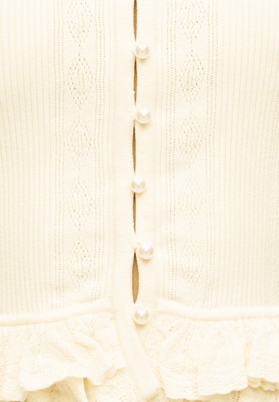 Κρεμ πλεκτή ζακέτα με κουμπιά από πέρλες, κατακόρυφα διαμάντια μοτίβα και λεπτομέρεια με βολάν στο τελείωμα.