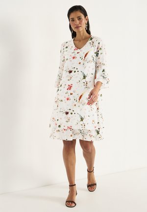 Vrouw loopt in een witte knielange jurk met bloemmotief, driekwart mouwen en zwarte sandalen met enkelbandje op een effen achtergrond.