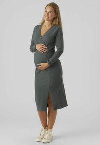 Zwangerschapsjurk in donkergrijs, lange mouwen, V-hals, gestructureerde stof, kniehoog met een zijsplit, gecombineerd met lichtgekleurde sneakers.