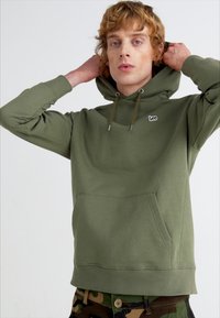 Felpa verde con tasca frontale, cappuccio con cordino e piccolo logo sul petto. Il tessuto sembra morbido con una trama semplice.