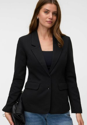 Kvinde iført sort blazer over sort top og blå jeans, der holder en sort foldet håndtaske mod en ensfarvet baggrund.