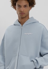 Felpa con zip di colore azzurro chiaro realizzata in tessuto morbido, con tasca frontale e logo "lowlights" ricamato in bianco sul petto.