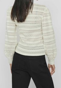 Pull en laine légère de couleur crème, tricoté, avec un motif rayé et des manches bouffantes. Porté avec un jean noir, mettant en valeur le tissu texturé et la coupe ajustée.