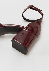 Sandalie cu toc înalt de culoare burgundă, cu finisaj lucios din patent, curea lată, vârf rotund și curea la gleznă ajustabilă cu cataramă metalică.