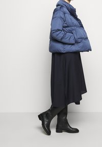 Ciemnoniebieska kurtka puchowa z wysokim kołnierzem, o szerokiej sylwetce i pikowanej teksturze, zestawiona z lejącą czarną spódnicą i czarnymi botkami za kolano.