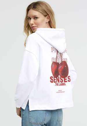 Woman with blonde hair wearing a white hoodie with red cherry graphic and French text, paired with blue jeans, facing backward and looking over shoulder.