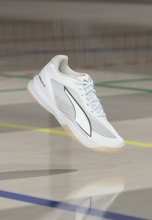 Chaussure de sport blanche Puma avec semelle beige, logo noir et lacets, flottant au-dessus d’un sol de gym marqué de lignes colorées.