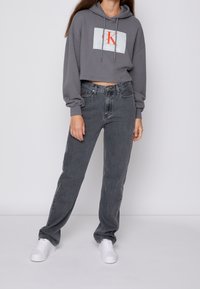 Kobieta ubrana w szary, krótki bluzę Calvin Klein, wysokie, wyblakłe czarne dżinsy i białe sneakersy, stojąca w białym tle.