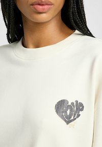 Kremowy sweatshirt z prążkowanym dekoltem i graficznym motywem serca w ciemnych paskach, z napisem "love" w środku.