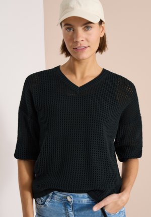 Femme portant une casquette blanche, un pull tricoté noir à manches courtes et un jean bleu, une main dans la poche sur fond beige.