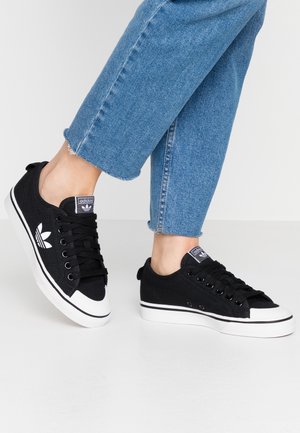 Osoba nosząca czarne tenisówki Adidas z białą podeszwą i logo, zestawione z podwiniętymi niebieskimi dżinsami na białej podłodze.