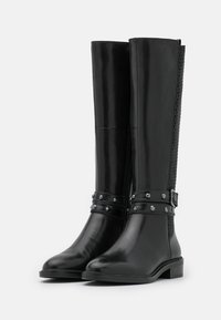 Bottes en cuir noir montant jusqu'aux genoux avec une finition lisse, un panneau latéral texturé, un détail de ceinture cloutée et un talon bloc bas.