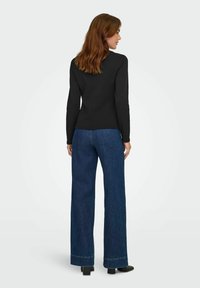 Zwart langslijp shirt gecombineerd met blauwe wijde jeans. Het shirt heeft een getailleerde pasvorm en de jeans hebben een rafelrand.