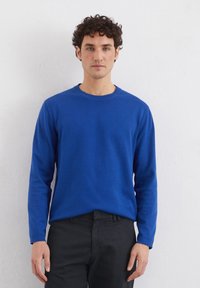 Blauer langärmeliger Baumwollpullover mit Rundhalsausschnitt und gerippten Bündchen, über dunklen Hosen getragen. Glatte Textur, einfaches Design.
