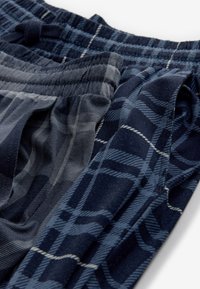 Close-up of three pairs of plaid flannel pajama pants in dark blue and gray tones with elastic waistbands and drawstrings.