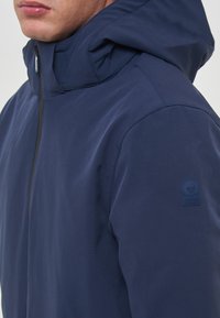 Veste en nylon bleu marine avec un col haut, une capuche et un patch logo circulaire sur la manche gauche. Texture lisse et design épuré.