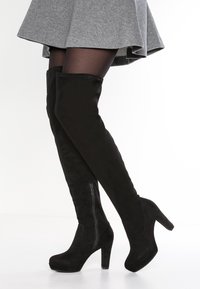 Botas de ante negras hasta la rodilla con tacón de aguja, que cuentan con un zipper lateral y textura suave. El eje de la bota es ajustado y elegante.