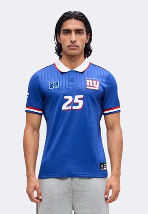 Homme aux cheveux mi-longs foncés portant un maillot de sport bleu avec un col blanc et le numéro 25, debout devant un fond uni.
