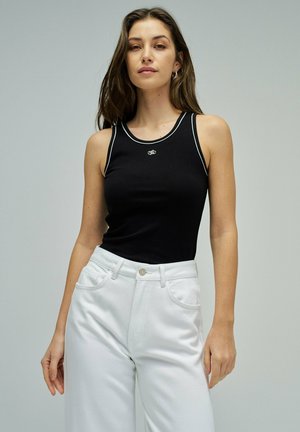 Jeune femme portant un haut noir sans manches et un pantalon blanc taille haute, debout devant un fond gris clair uni.