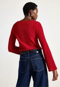 Rød tekstureret langærmet top med et bølget mønster, parret med høj talje mørke denim jeans med to baglommer og et sort bælte.