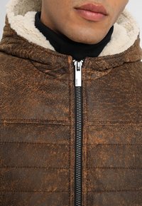 Muž v hnědé strukturované zipové bundě s kapucí podšitou sherpau a černém roláku uvnitř, zobrazen od brady po hruď.