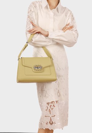 Donna che indossa un vestito e una camicetta bianchi in pizzo, mentre tiene davanti a sé una borsa a mano in pelle verde chiaro con una chiusura metallica.