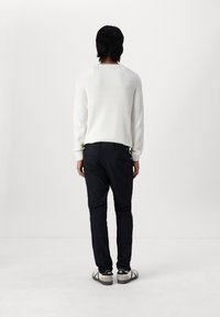 Vit ribbad tröja, marinblå byxor och vita skor med svarta detaljer. Personen visas bakifrån, vänd mot en minimalistisk bakgrund.