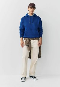Blaues Hoodie mit Kordelzug, beige Hose und ein kariertes Hemd um die Taille gebunden. Schwarze Sneakers runden das Outfit ab.