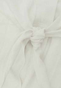 Nahaufnahme eines weißen Stoffgürtels, der in der Mitte eines Kleidungsstücks mit sichtbaren Nähdetails zu einem Knoten gebunden ist.