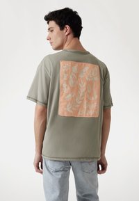 Oliven grøn bomulds t-shirt med en firkantet lys ferskenfarvet grafik på ryggen, der har hvide botaniske mønstre og tekst, der siger "Guess Los Angeles."