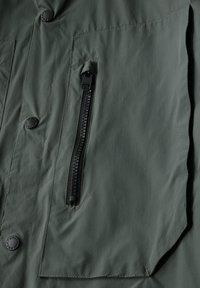 Zelená vnější látka s dvěma kapsami s klopami, zipovou kapsou s černými kovovými doplňky a hladkým, mírně texturovaným povrchem.